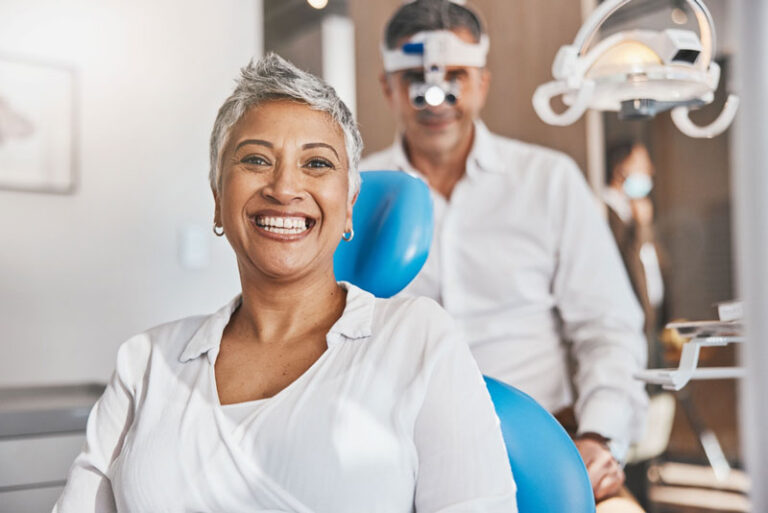 Smiling dental patient sitting in a treatment chair with a dentist in the background wearing magnifying loupes in a modern dental clinic.